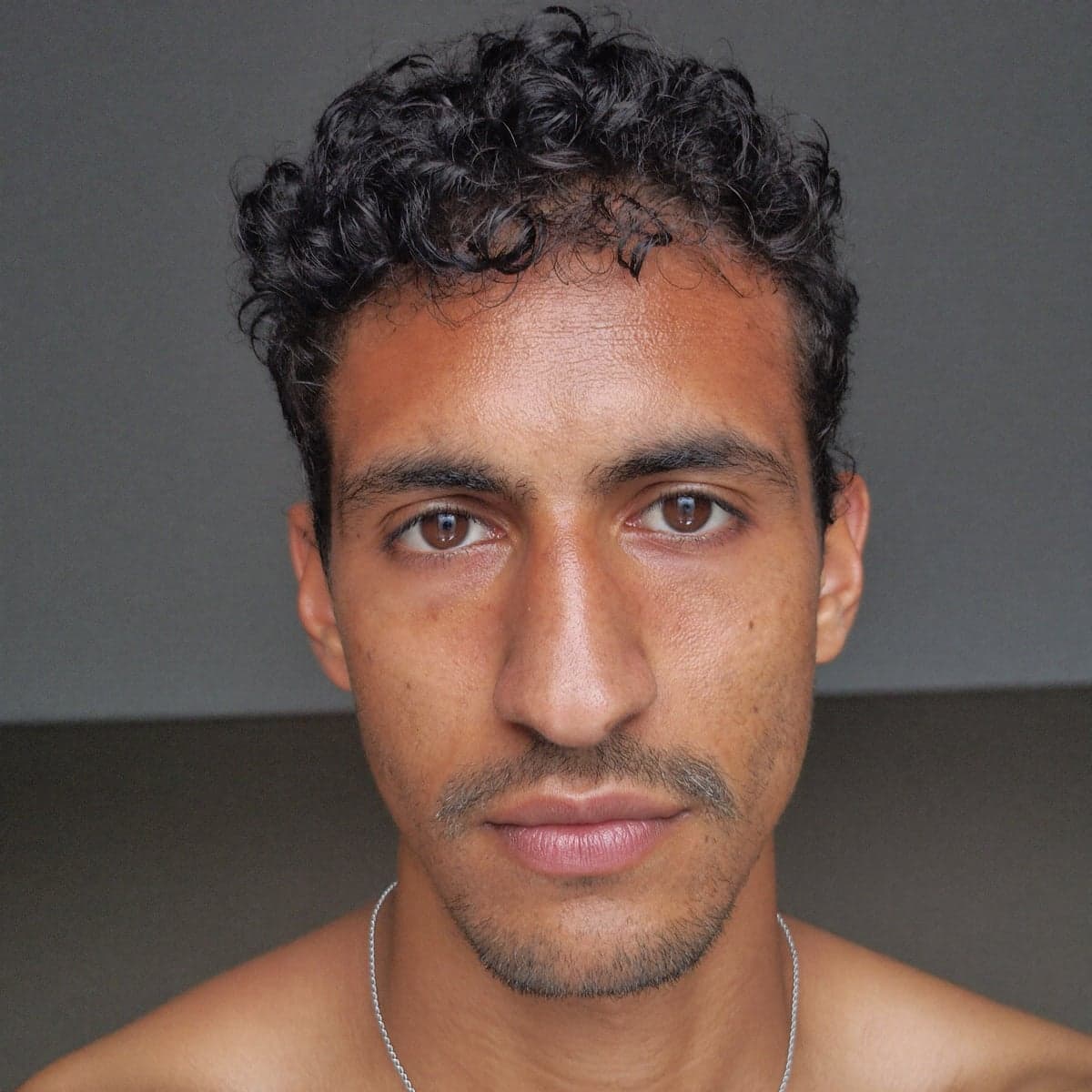 Phone headshot of a man with dark curly hair against a grey wall