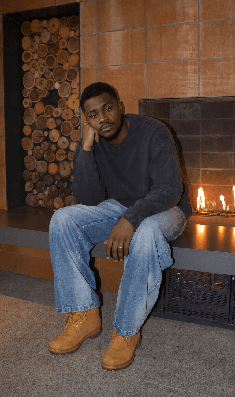 Man in a navy sweater and jeans sitting by a fireplace